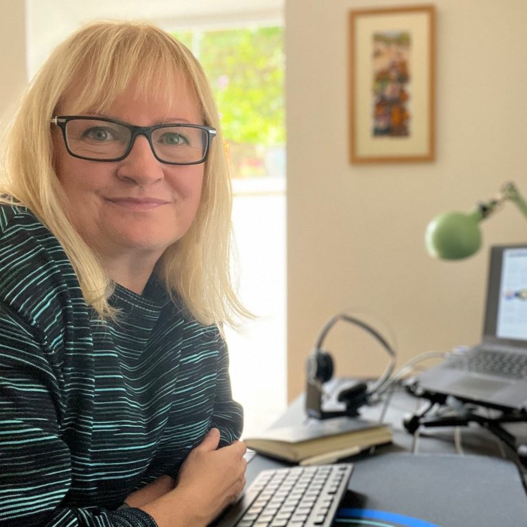 White woman with blonde hair wearing black/green jumper at a desk with keyboard and headphones