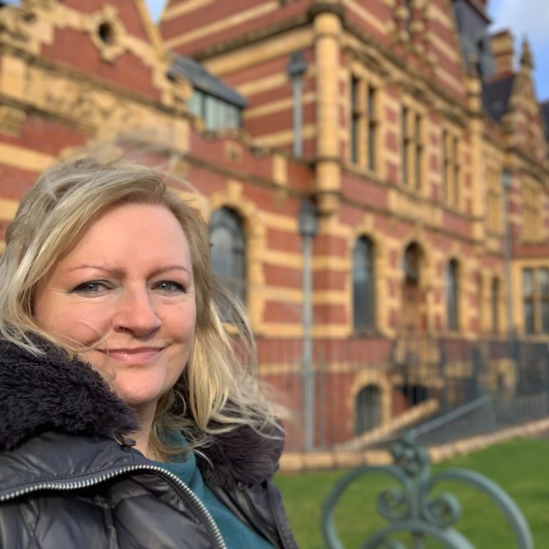 Smiling white woman with black rimmed glasses and pink jumper overlooking a heritage swimming pool filled with people and inflatables